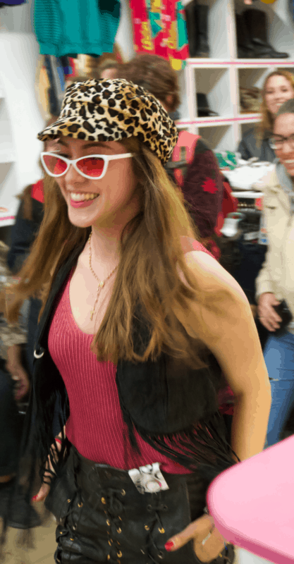 Picture shows model wearing leopard print hat, white rimmed sunglasses with pink lenses, pink knitted vest top, leather waist coat with tassels and leather trousers with lace up detail. 